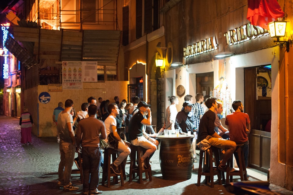 Посетители за столиками-бочками возле пивного паба Birreria Trilussa вечером в Trastevere, Рим